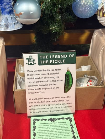 The image shows a Christmas ornament set featuring "The Legend of the Pickle". The display includes a card explaining the tradition: German families place a pickle ornament on the Christmas tree last. On Christmas Day, the child who finds the pickle first receives an extra gift from St. Nicholas for being observant. The set also contains a pickle-shaped ornament. A second card elaborates on the tradition of the pickle ornament.
