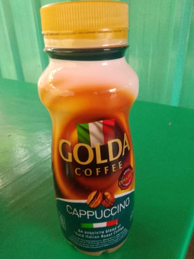 A plastic bottle of Golda Coffee cappuccino is placed on a green surface. The bottle has a yellow cap and a label with the Golda Coffee logo, an Italian flag, and the word "CAPPUCCINO" printed in large white letters. There is also text in smaller font on the label, and an image of two coffee beans. The background is a blurry green texture, possibly a wall or curtain. The lighting is direct, causing reflections on the bottle. The scene appears to be indoors. No people or specific location in Bogor, Indonesia are visible in the image.