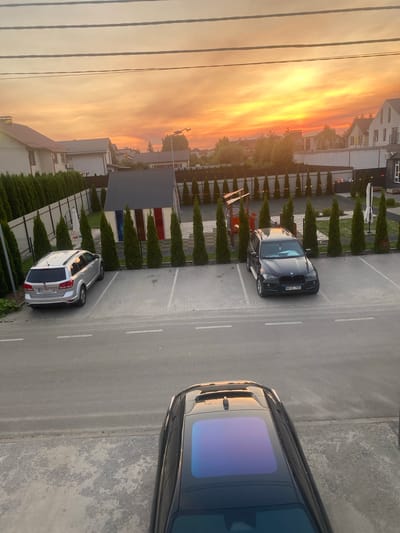 The image depicts an outdoor parking lot bathed in the warm light of a sunset in Novi Petrivtsi, Ukraine. A black SUV with a sunroof is parked in the foreground, facing away from the viewer. In the middle ground, a silver SUV is parked to the left, and a black BMW SUV is parked to the right. Behind the vehicles, a row of manicured evergreen trees lines a fence. Further back, a small building with a gray roof and colorful panels is visible, along with more evergreen trees and residential buildings. The sky is filled with vibrant orange and yellow hues, indicating either a sunrise or sunset. The ground is paved with interlocking bricks in the parking area, transitioning to asphalt on the road. There are no people visible in the image.