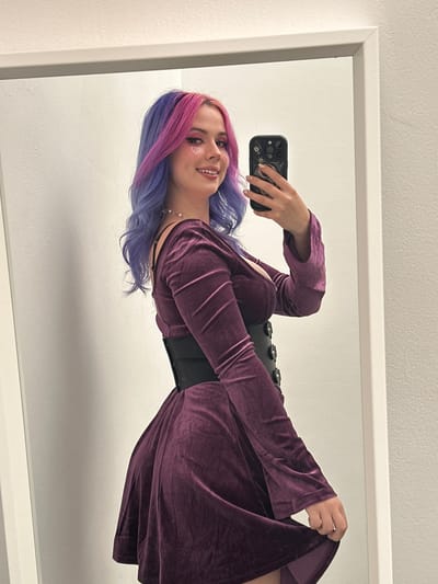 A woman is posing for a mirror selfie. She is wearing a long-sleeved, purple velvet dress and a wide black corset. Her hair is dyed with purple and blue streaks, and she has heart-shaped glitter stickers on her face. She is holding a black smartphone in her right hand, and the phone case has a detailed design. The mirror has a white frame. The wall behind the mirror is white. The location is not discernible from the image.