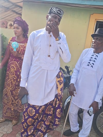 The image depicts three individuals in Rido, Nigeria. An older man wearing a white traditional Igbo attire with black embellishments, a black fedora hat, and black shoes is partially visible at the top. He is holding a walking stick and what appears to be a handkerchief or small cloth. Below him, a man in a white Igbo-style shirt with brown and gold embellishments and a dark-colored cap with white embroidery is seen covering his mouth with his hand. He wears a gold chain and a watch. He also wears a dark-patterned fabric wrapped around his waist. At the bottom of the image, a woman in a purple headscarf and a purple and gold embroidered dress is holding a small blue fan. The background shows a light yellow wall. They stand on a tiled pavement. The image seems to have been taken during a sunny day.