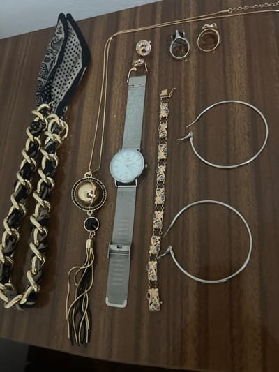 Multiple items of jewelry and accessories are arranged on a polished, medium-brown wooden surface, exhibiting a vertical wood grain pattern. The scene is located in the city of Smederevska Palanka Municipality, Serbia.

At the top left, a folded scarf, predominantly black and off-white with a patterned design, is positioned, attached to a thick, gold-colored chain interwoven with dark fabric. Below this, a long, gold-colored necklace features a circular pendant with a central gold sphere, a ring of small black beads, and a hanging black stone from which gold and black tassels extend.

Centrally placed is a silver-colored wristwatch with a white face displaying "COLMAR" and an unclasped silver mesh strap. To its right, an unclasped gold-colored bracelet features alternating rectangular and oval links, with the rectangular links inlaid with small blue or purple stones.

At the top right, a thin, gold-colored chain necklace is laid horizontally. Below it, from left to right, are a small gold-colored stud earring with a pink stone, a silver-colored ring with a dark stone set in a crown-like bezel, and a plain gold-colored band ring.

Two large, unfastened silver-colored hoop earrings are positioned vertically stacked on the bottom right of the frame.