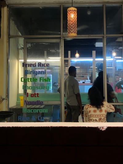 The image depicts the interior of a restaurant in Ahangama, Sri Lanka, viewed through a large glass window. The window displays a list of food items in colorful lettering on the glass, including "Fried Rice," "Biriyani," "Cuttle Fish," "Chopsuey," "Lott," "Noodle," "Macaroni," and "Rice & Cu." Inside the restaurant, several individuals are visible. A man in a gray t-shirt and brown pants stands facing away from the camera, looking into the restaurant. A woman with long, dark curly hair is seated facing the man, wearing a patterned white top with red and yellow sun motifs. Another woman, partially obscured, is visible behind her. In the background, another man in a pink shirt is partially visible, along with a refrigerator with "Pepsi" branding. Fluorescent lights are visible above the counter, and a decorative woven light fixture hangs from the ceiling. The overall scene suggests a casual dining establishment.