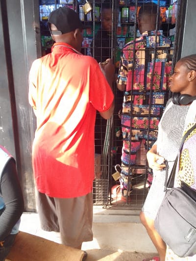 The scene depicts an outdoor commercial interaction in Nkwelle Ezunaka, Nigeria. Three individuals are prominently visible, with two others partially obscured. In the foreground, a male subject is seen from behind, wearing a bright red polo shirt, dark shorts, and a black baseball cap reversed; he appears to be reaching towards a black metal security gate. To his right, a female subject stands in profile, facing left, wearing a light patterned dress and black over-ear headphones around her neck, carrying a black shoulder bag. Behind the security gate, secured by a brass-colored padlock, a second male subject, also seen from behind, wears a distinct multicolored patterned shirt. He stands within what appears to be a stall or shop, densely packed with various electronic accessories, likely for mobile phones, displayed on shelves. A third individual is partially visible behind him, wearing dark attire. In the extreme lower-left, a portion of a person in blue and black clothing is visible next to a flat wooden surface on the light-colored ground. The scene is illuminated by strong overhead sunlight, casting defined shadows.