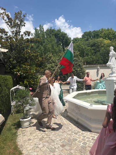 A group of people are gathered outdoors, possibly for a celebration or wedding, in Haskovo, Bulgaria. The scene is set in a garden with lush green trees and a cobblestone pathway. A white fountain with water is a prominent feature.
Several individuals are interacting, with some appearing to dance. One person is holding a Bulgarian flag, suggesting a national celebration or pride. Another individual is dressed in a wedding-like attire, hinting at a wedding event. The sky is a clear blue with some white clouds, indicating a pleasant day, likely daytime. The overall mood seems joyful and celebratory, with people holding hands and smiling.