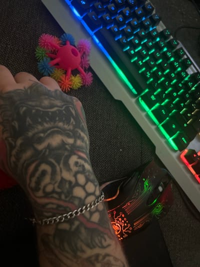 The image shows a close-up view of a person's hand with a tattoo, resting on a textured surface, possibly a desk or a couch. To the right of the hand is a white gaming keyboard with colorful LED backlighting in blue, green, and red. In front of the keyboard is a black gaming mouse with a circuit board design illuminated by red and green LEDs. To the left of the keyboard, near the hand, is a small pink toy with multiple colorful fuzzy attachments. The overall scene suggests a personal workspace, likely for gaming or computer use. There is no identifiable location within the city of Gelsenkirchen, Germany.