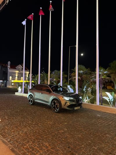This is a nighttime, eye-level, outdoor photograph taken in Cascais, Portugal. The scene features a silver crossover SUV parked on a cobblestone driveway, illuminated by artificial lights. In the background, a building with illuminated windows is visible, along with several tall flagpoles with flags of various colors, including red, white, and dark red. Palm trees and other tropical foliage are also present, adding to the ambiance. The moon is visible in the dark night sky. The car has its headlights on, and a license plate is visible on the front of the vehicle. The lighting suggests it is likely evening or night.