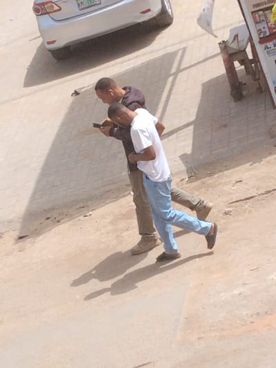 An elevated view captures two adult males walking on an unpaved, light brown surface in Fagge A, Nigeria, during daylight hours. The male in the rear wears a dark long-sleeved shirt, khaki pants, and light brown shoes, holding a smartphone in his right hand and looking at its screen. White earbuds are visible in his ears. The male in front wears a white short-sleeved t-shirt, light blue pants, and dark open-toed sandals, also directing his gaze towards the smartphone held by the other individual. Both men exhibit closely cropped dark hair and are casting distinct shadows on the ground, indicating strong overhead light. In the upper left background, a silver sedan, likely a Toyota, is partially visible on a paved surface, displaying a Nigerian license plate with green characters, partially reading "KMC 793G". To the far right, a portion of a vendor's stall or sign structure, with items displayed and a white fabric piece attached, is discernible.