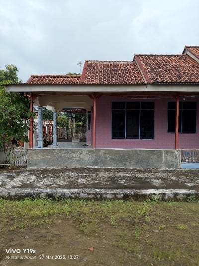 The image shows the front of a single-story house in Anjatan, Indonesia, located at coordinates -6.419 latitude and 107.978 longitude. The house has a red tile roof and a pink stucco exterior. There is a covered porch with four white pillars that extends across the front of the house. The porch is empty. The house has a window on each side of the porch. The window on the left side of the porch is large and consists of six panes of glass, while the window on the right is smaller with only three panes of glass. There is a wooden fence and some trees in front of the house. The ground in front of the house is covered in dirt and grass. There are no people in the image.