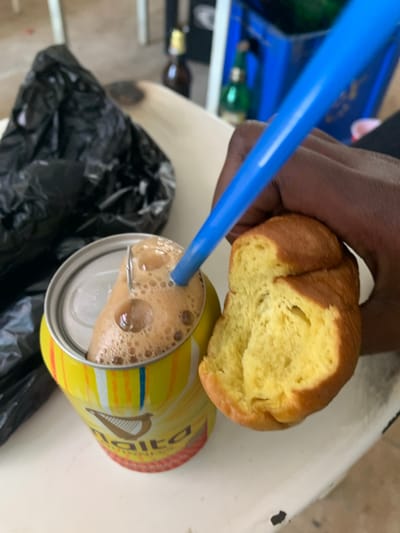 In New Stadium, Nigeria, a person's hand is holding a piece of bread over a can of Malta Guinness. The can is yellow with red and blue stripes and features the Guinness harp logo. A blue straw is inserted into the can, and the drink is frothy. A black plastic bag is visible in the background, as are some green bottles and blue crates. The scene appears to be outdoors, possibly on a table.