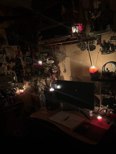 The image depicts a dimly lit, cluttered room in Vladaya, Bulgaria, possibly a workshop or storage area. The overall impression is one of disarray and perhaps abandonment, with various objects strewn about. Prominent subjects include what appear to be decorative elements and potentially discarded items. There is a red lantern with a light on, casting a warm glow, and another red light emitting from a crystal-like object on a desk. A television screen is visible, though it is dark, and a stack of papers with some writing on them rests on the desk. Other objects scattered around include what look like headphones, car emblems, a plastic water bottle, and what could be toy weapons or tools. The lighting suggests it is nighttime or a very dark indoor space. There are no people visible in the image. The clutter and dim lighting create a somewhat mysterious or melancholic atmosphere.