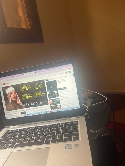 The image shows a laptop on a desk, displaying a YouTube video of Quranic recitations. The video features Arabic script and images of a reciter. The laptop's keyboard is visible, and headphones are plugged in nearby. The background includes a wall, a window frame, and a section of what appears to be a patterned surface. The video title displayed on the screen is "Listen with the intention of Rizk (sustenance) and healing." The image appears to have been taken indoors. The OCR text hints at location of Morocco, since "MA" is at the bottom left. No evidence indicates the specific city of Ouaouizert.