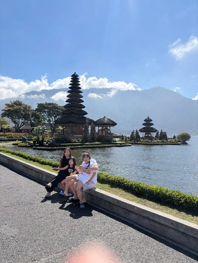 A medium shot captures four people sitting on a concrete ledge with a scenic background of Pura Ulun Danu Bratan temple complex in Baturiti, Indonesia. The temple features a multi-tiered pagoda structure situated on an island in the middle of a calm lake. Mountains and clouds form the backdrop. Two women and two children are seated on the ledge. The woman on the left wears a black dress and black pants. Beside her, a young girl in a black t-shirt and light-colored shorts is held by another woman wearing a white t-shirt and white shorts. Another young girl in a white shirt and shorts is seated on the woman's lap. All are looking towards the temple complex. The lake is tranquil, reflecting the blue sky and the temple structures. The foreground shows a paved path and a grassy area bordering the lake.