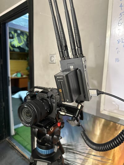 The image captures a detailed view of a professional video camera setup, likely in a studio or production environment in Ikeja, Nigeria. The primary subject is a mirrorless camera, specifically a Panasonic Lumix, equipped with a Panasonic Lumix S 20-60mm f/3.5-5.6 lens, visible from a downward angle. The camera is enclosed within a protective cage or rig, with various mounting points and accessories attached.
Mounted above the camera is a wireless video transmitter/receiver unit, distinguishable by its gray metallic housing and three prominent external antennas. A Sony NP-F970 InfoLITHIUM L battery powers this unit, directly attached to its side. A black USB cable is plugged into the top of the wireless unit, connecting it to an unidentifiable device. Another coiled black cable extends from the camera rig upwards, suggesting a power or video connection.
In the background, a white whiteboard is visible with handwritten numbers "1 2 3 4 5 6 7 8 9 10" in blue marker. Reflections on a surface below the camera reveal a blurred indoor scene, including glimpses of furniture and possibly other objects. The setup indicates active use or preparation for video recording or live production, utilizing wireless video transmission for monitoring.