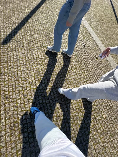 Three individuals are observed on a cobblestone pavement characteristic of Portuguese cities, likely in Bragança, Portugal. The ground is composed of small grey stone blocks, with visible green moss in the interstices. A faint white line traverses the pavement. Strong, elongated shadows are cast on the ground, indicating direct sunlight from a low angle.
The first individual, positioned towards the upper left, is seen from the mid-thigh down, wearing light blue, loose-fitting jeans and white athletic shoes with dark accents. A grey garment is visible above the waistline.
The second individual, located towards the mid-right, is also seen from the mid-thigh down, dressed in light grey sweatpants and white athletic shoes. This person holds a white smartphone with a purple-patterned screen in their right hand, oriented away from the viewer.
The third individual, whose lower legs and feet are visible at the bottom-left, appears to be the photographer. This person wears light-colored (possibly white or light blue) pants and blue athletic shoes with black detailing.
The individuals are in close proximity, standing or walking on the sunlit pavement. The specific location within Bragança cannot be precisely identified from the image content beyond being a public outdoor area with traditional Portuguese pavement.