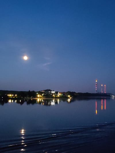 The image depicts a twilight scene of the Volga River in Konakovo, Russia. In the distance, the riverbank is illuminated by the lights of buildings and industrial smokestacks, which reflect on the water's surface. The moon is visible in the clear, dark blue sky, casting a soft glow. The foreground shows ripples on the water, indicating movement. There are no people visible in the frame. The overall atmosphere is serene and tranquil, capturing the quiet beauty of the waterfront at night.