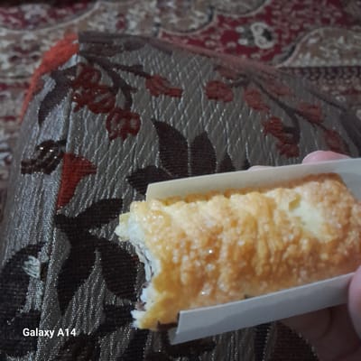 A close-up, low-angle shot shows a hand holding a pastry with a bite taken out of it, against a patterned textile background. The pastry is golden brown and appears to be dusted with sugar. The textile has a dark brown base with a textured, repeating pattern of stylized red floral motifs and darker brown leaf shapes. The word "Galaxy A14" is visible in white text in the lower left corner of the image. The scene is indoors, likely in a home environment in Khan Yunis, Palestinian Territories, given the watermark.