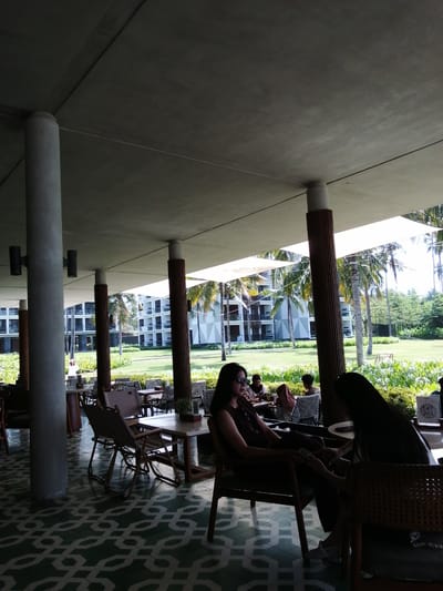 A low-angle, medium shot captures an outdoor seating area with a thatched roof supported by concrete pillars and wooden posts, casting shadows on the patterned floor. In the foreground, two women are seated at a table, with the woman on the left wearing sunglasses and a dark sleeveless top, holding a phone. The woman on the right, in silhouette, has long dark hair and appears to be interacting with the first woman. In the background, several other patrons are seated at tables, with lush greenery, palm trees, and a modern white building visible beyond. The scene suggests a resort or hotel setting. The location is identified as Banyuwangi, Indonesia, likely a resort with an outdoor cafe or dining area.