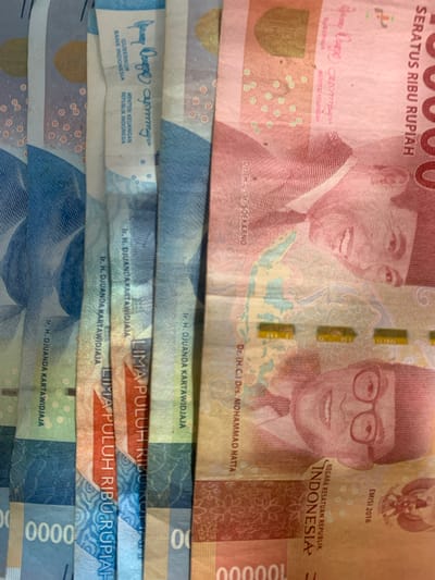 This media file displays a close-up, high-angle view of stacked Indonesian rupiah banknotes. The primary subjects are the portraits of Indonesian historical figures featured on the currency. On the top banknote, a red one, two men are visible: Soekarno on the left and Mohammad Hatta on the right. The text "SERATUS RIBU RUPIAH" indicates the denomination of 100,000 Rupiah. Below the portraits, the names "Dr. H.C. Ir. SOEKARNO" and "Dr. (H.C.) Drs. MOHAMMAD HATTA" are visible. The emission year "EMISI 2016" is also present, along with "NEGARA KESATUAN REPUBLIK INDONESIA".
Below the top banknote, several other denominations are partially visible. These include banknotes with blue and green color schemes. Textual elements on these lower banknotes include "Ir. H. DJUANDA KARTAWIDJAJA", "GUBERNUR BANK INDONESIA", and "MENTERI KEUANGAN REPUBLIK INDONESIA". The phrase "LIMA PULUH RIBU RUPIAH" (Fifty Thousand Rupiah) is also clearly legible on some of these.
The lighting suggests an indoor setting or possibly an overcast day, as there are no strong shadows. The focus is on the details of the banknotes, highlighting their design and text. The image's composition, with the overlapping bills, creates a sense of abundance or a collection of money. There are no overt emotional cues, but the image is informative in its display of currency. The specific location mentioned as Sungai Meriam, Indonesia, provides a geographical context for the currency being Indonesian.
