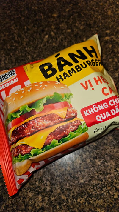 The image shows a close-up of a package of "Bánh Hamburger" flavored crackers. The package is predominantly yellow, red, and white, with a picture of a cheeseburger. Text in Vietnamese and Chinese is visible on the packaging. The background is a dark, speckled surface, possibly a countertop. The crackers are most likely in Cam Duc, Vietnam.