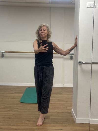A person, appearing to be female, is seen in what appears to be a selfie taken in a room. The person is light-skinned with wavy, blonde/gray hair, and is wearing a black tank top and dark patterned leggings. She is holding a black smartphone in her left hand, presumably taking the photo. Her right arm is extended, touching the upper wall of the room. The environment is a room with white walls, a wooden floor, and a ballet barre on the wall. A green yoga mat lies on the floor. The location in Ramat Gan, Israel, is indeterminable.