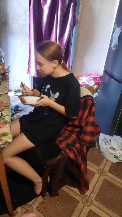 The image shows a young woman sitting on a chair, eating from a bowl. She is wearing a black t-shirt and shorts. A red and black plaid jacket is draped over the back of the chair. The room appears to be a kitchen or dining area, with a refrigerator in the background. The floor is tiled with a brown and beige pattern. There are curtains in the background, a table with a floral tablecloth on the left, and various items on the floor. The scene suggests a casual domestic setting, possibly in an apartment or house. There are no visible indicators in the image to suggest the specific location in the city of Samar, Ukraine.