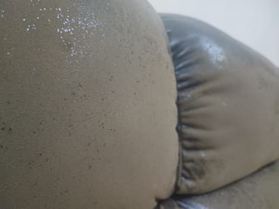 The image shows a close-up, abstract view of a textured surface, likely upholstery from a piece of furniture. The dominant color is a muted, grayish-brown. The surface on the left is lighter and speckled with small, reflective white flecks, resembling glitter or a metallic thread woven into the fabric. There are also scattered dark specks, suggesting dirt or wear. To the right, another portion of the surface is visible, this one appearing darker and shinier, with visible creases and folds. The lighting in the image creates highlights and shadows, emphasizing the texture and curvature of the material. The overall impression is one of a worn or aged fabric, possibly from a sofa or chair. There are no discernible elements that would indicate a specific location in Pattaya City, Thailand, or any people, actions, or interactions.