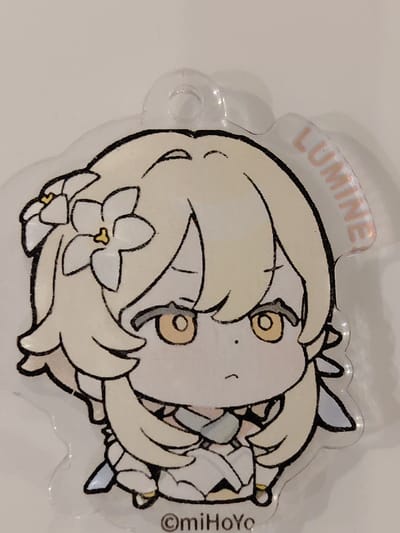 This media file features a close-up of a clear acrylic keychain with a character illustration. The character is drawn in a chibi anime style, with a large head and simplified features. She has light blonde hair adorned with white flowers, and large golden eyes with dark pupils. Her expression appears neutral or slightly downcast, with a small frown. She is wearing a white dress with light blue accents. The keychain has a hole at the top for attachment. The background is a plain, light-colored surface, likely a table or wall.

Visible text on the keychain includes the name "LUMINNE" printed in a light pink, curved font along the upper right edge. At the bottom center, there is a copyright symbol followed by "miHoYo", indicating the character or product is associated with the company miHoYo, known for creating video games such as Genshin Impact, from which this character likely originates. The overall presentation suggests a merchandise item related to a popular game or media franchise. The lighting is even, with no strong shadows, suggesting indoor lighting. The location context of Balashikha, Russia, does not offer any visual cues from the image itself.