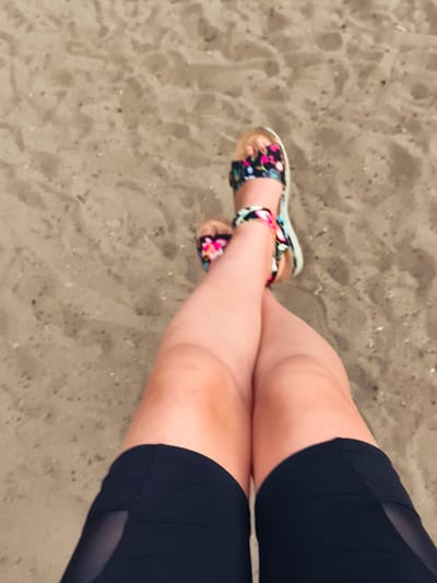 The image shows a person's legs crossed, resting on sand. The person is wearing black shorts with mesh detailing and floral-patterned sandals. The setting appears to be a beach. There is no specific indication of location within the city of Wrocław, Poland, as the image only shows the legs and sand.