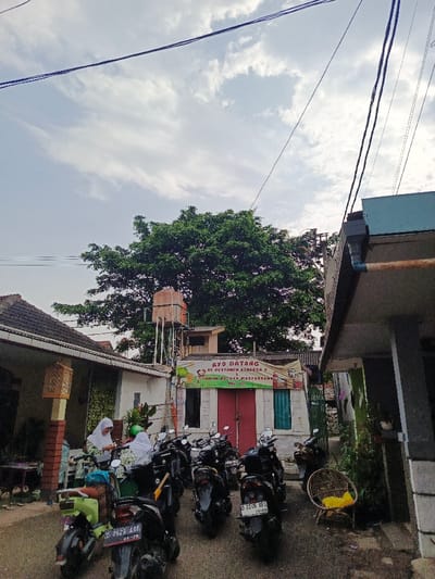 A street scene shows several parked motorcycles facing a building with red doors. A sign above the doors reads "AYO DATANG KE POSYANDO KENANGA 2" and includes other text. Two women in white headscarves are near a green scooter. A water tank is visible behind the building, and a large tree looms in the background under a cloudy sky. Power lines stretch across the top of the image. The area appears residential with houses on either side of the street.