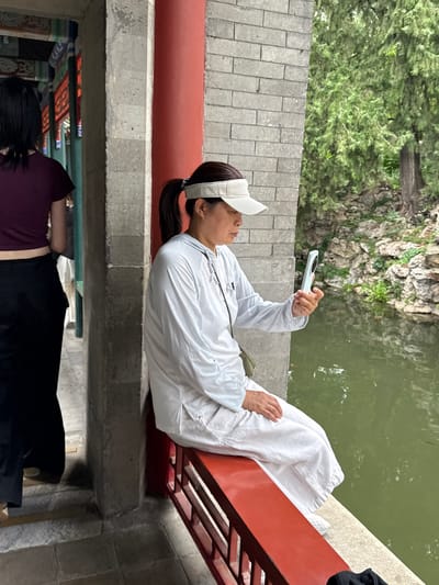 This image depicts a woman sitting on a red railing, holding a light blue smartphone and looking at its screen. She is wearing a white long-sleeved shirt, white pants, and a white visor. To her left, another person is visible from the back, wearing a maroon shirt and black pants, standing in an indoor area with decorative beams. The background features a body of water and lush green trees. The architecture suggests a traditional Chinese setting, likely within Xicheng District, China, possibly a historical site or park. The woman is positioned on an elevated walkway or bridge overlooking the water, which appears to be a pond or canal. The stone wall to her right indicates a robust, possibly ancient, structure. The lighting suggests an overcast day.