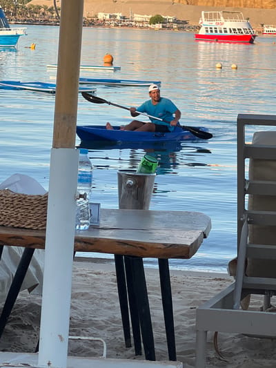 The image shows a man in a blue shirt and white cap kayaking on calm, blue water. He is holding a black paddle and is positioned in the middle of a blue kayak. The kayak is on the water, which reflects the sky and a distant boat. In the background, a red and white boat with "FUN DAY" written on its side is visible, as are several buoys. The scene appears to be a beach or waterfront area, with sand and some structures visible on the shore. A wooden table and chairs are in the foreground, suggesting a seating area near the water. There is a green bottle and two bottles of water on the table. The lighting suggests it is daytime, likely morning or late afternoon given the warm tones in the water. The overall mood is relaxed and recreational. The location is Sharm El Sheikh, Egypt.