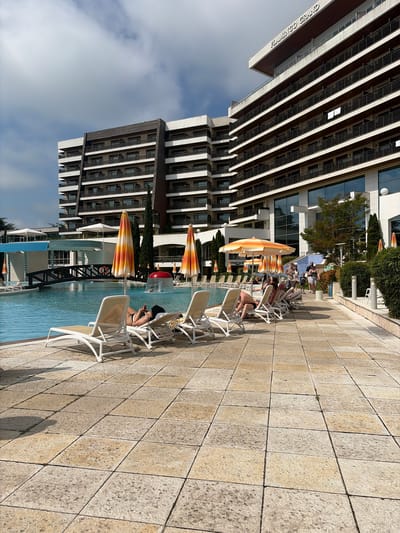 The image captures an outdoor scene at the Flamingo Grand hotel resort in Obrochishte, Bulgaria. The multi-story hotel building, visible prominently in the background and upper right, features a modern design with light-colored facades, dark brown accents on balconies, and expansive glass windows reflecting the sky and surroundings. A second, similar architectural building is partially visible to the left.
In the midground and foreground, a large, irregularly shaped swimming pool with clear blue water is present. An arched, dark-colored bridge spans a section of the pool, with a red life preserver hanging from its railing. A red, mushroom-shaped water feature actively sprays water into the pool.
The poolside patio is paved with large, light-colored square tiles. A row of white sun loungers are arranged on this patio; several individuals are relaxing on them. One woman in a black and white bikini is lying on a lounger, looking at a mobile phone. Multiple large, striped parasols in shades of orange, yellow, and white are interspersed around the pool, providing shade.
A paved pathway runs parallel to the pool on the right, where several people are walking casually. This group includes adults and a partially visible stroller. Green hedges line this path, and tall, slender evergreen trees are visible throughout the resort grounds. Signs indicating "NIGHT CLUB" and "EXIT" are visible near the umbrellas and pathway. The sky above is partly cloudy, with patches of blue visible.