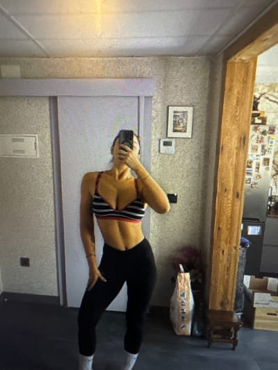 In this full-body, mirror-reflected image, a person is standing in what appears to be a residential interior. The individual is wearing a black sports bra with horizontal white stripes and red trim, paired with black leggings and white socks. They are holding a smartphone to take the picture, obscuring their face. The background features a light-colored wall with textured wallpaper, a sliding door to the left, and a wooden post on the right. Several framed pictures and what looks like a refrigerator with items attached to it are visible in the background. The floor is a dark color, possibly a mat or dark tile. The overall impression is of a personal space, possibly a bedroom or hallway, where a fitness-related photograph is being taken. There are no discernible elements in the image that suggest a connection to the city of Murcia, Spain.