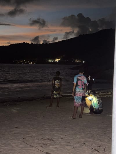 The image depicts a nighttime scene at a beach in Rawai, Thailand. Four individuals are present. In the foreground, a person stands facing away from the camera, wearing a black t-shirt with an eagle graphic and text that appears to be "BORN TO BE WILD" and "ARMY AIR CORP". They are also wearing patterned shorts.
Further back, two individuals are bent over, seemingly interacting with a mobile device. One wears a t-shirt with the number "56" in large pink numerals on a blue and pink patterned background, paired with matching shorts. The other person is wearing a t-shirt with "56" in white numerals on a green and yellow gradient, with black shorts.
A fourth person is partially visible in the background, hunched over a laptop.
The setting is a sandy beach next to a body of water, likely the ocean. Distant lights suggest a coastal town or resort. The sky shows remnants of twilight with clouds in shades of gray and faint orange, indicating the time of day is either late dusk or early dawn. The overall atmosphere is relaxed and casual, possibly a group enjoying a quiet evening by the sea.