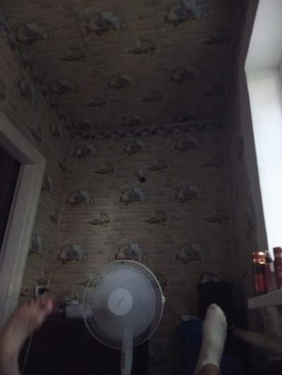 The media file shows a dimly lit interior scene, likely a room in a residential building in Yenakiieve, Ukraine. The walls and ceiling are covered in wallpaper with a repeating pattern that appears to be stylized representations of clouds and brickwork. A white oscillating fan stands in the foreground to the right of the image. To the left, a person's legs and feet are visible, with one foot wearing a white sock. The lighting suggests it is either nighttime or the room has limited natural light, with a window visible on the far right. The overall atmosphere is ordinary and domestic. There is no visible text in the media file.