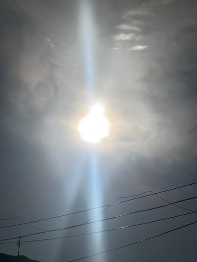 The image shows a bright, hazy sun partially obscured by clouds. A strong beam of light emanates from the sun, creating a lens flare effect. In the foreground, several power lines are visible, stretching across the frame against the cloudy sky. The scene appears to be outdoors, with a hint of land visible in the lower left corner, suggesting a potentially elevated viewpoint. The overall impression is one of a bright, overcast day. The location is identified as Abovyan, Armenia.