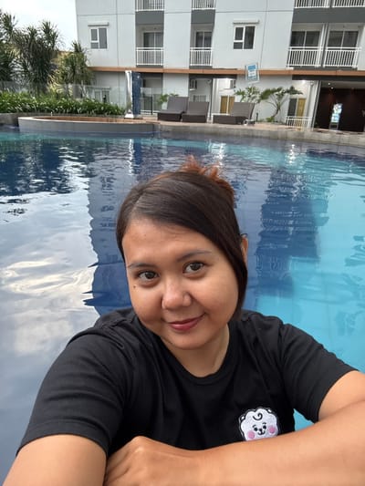 This image captures an adult female at the edge of a swimming pool in Sidoarjo, Indonesia. The subject, positioned in the foreground and slightly right of center, has dark brown hair pulled back and wears a black short-sleeved t-shirt with a white and pink cartoon character embroidered on the left chest. She faces the camera directly, displaying a subtle smile, with her forearms resting on an unseen pool edge. The midground features a blue swimming pool, reflecting an overcast sky and elements of the background structure. Behind the subject within the pool area, a curved, raised concrete feature is visible, possibly part of a jacuzzi. The background is dominated by a multi-story, light-colored modern building, characterized by numerous windows and balconies with white railings, indicative of an apartment complex or hotel. Adjacent to the pool on a paved deck are several dark brown lounge chairs, interspersed with green foliage including shrubs and small palm trees. A silver-toned artistic sculpture and a blue informational sign are also visible near the building by the pool. The scene is illuminated by natural daylight.