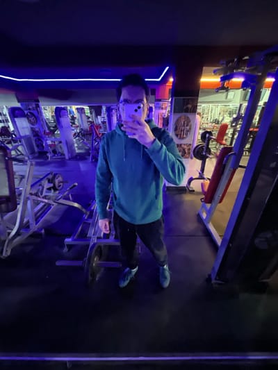 The image displays the interior of a gym located in Fez, Morocco. A single individual stands centrally, captured in a mirror selfie. The person wears a teal-colored hooded sweatshirt, dark sweatpants, and blue athletic shoes. They are holding a light purple smartphone, which obscures the majority of their face; only eyeglasses are visible above the phone. The gym features dark flooring, likely rubber, and a ceiling illuminated by prominent blue neon strip lighting, casting a distinct blue ambient light throughout the space. Some areas in the background exhibit orange lighting. Various pieces of gym equipment are visible, including multiple leg press machines, weight racks, a barbell with weight plates resting on the floor near the subject's feet, and a red-cushioned weight bench positioned to the right. Walls are lined with mirrors, reflecting additional gym machinery and extending the visual depth of the room.