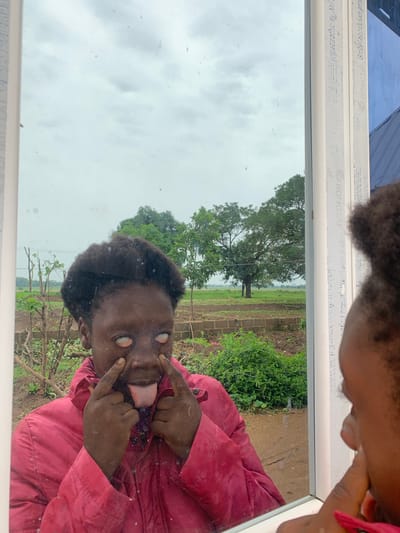 A photograph taken in Agban, Nigeria, shows a dark-skinned individual in a red jacket positioned at an angle in front of a mirror, pulling their eyes open with their fingers and sticking out their tongue. The reflection reveals the individual and a background featuring a grassy area with a large tree, possibly indicating a rural or outdoor setting. The sky is overcast. The mirror has white framing with some writing along the frame.