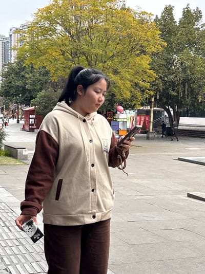A young female subject is depicted standing outdoors in an urban public space within Shapingba District, China. The subject, appearing East Asian, is positioned slightly left of center, viewing the screen of a black smartphone held in her right hand. A braided brown and red strap is attached to the phone. In her left hand, she holds a white and black cigarette pack featuring a panda design and Chinese characters. She wears a beige hooded jacket with contrasting dark brown sleeves and black buttons, along with dark brown corduroy pants. Her dark hair is pulled into a high ponytail with a white elastic band.

The background reveals a bustling urban environment under an overcast sky. To the subject's left, a pedestrian walkway is visible with small red kiosks, benches, and multiple distant figures. Tall, light-colored multi-story buildings rise on the far left. A large deciduous tree with prominent yellow-green foliage stands in the mid-ground, indicating autumn. Adjacent to it on the right is another large tree with darker green leaves. Further right, a colorful, cube-shaped art installation with a pink balloon attached is present. In the distant right background, two male figures are visible near a multi-tiered stone structure; one individual appears to be manipulating a wheeled cart. The ground is paved with light grey rectangular tiles.