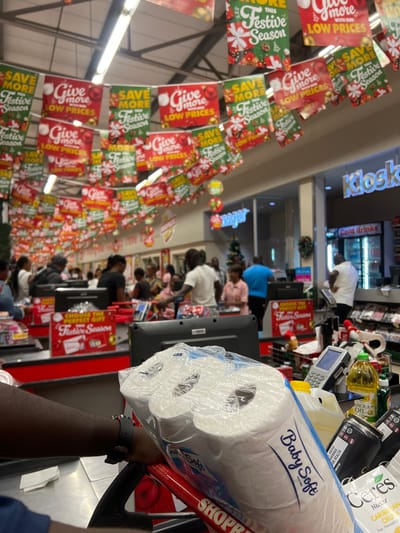 This image captures a bustling supermarket checkout area in Lusaka, Zambia, during the festive season. The perspective is from a customer's shopping cart, looking forward down a row of multiple active checkout lanes.

In the immediate foreground, a clear plastic-wrapped package of "Baby Soft" toilet paper rests on what appears to be a red shopping cart handle, with the partial brand name "SHOPR" visible, likely indicating Shoprite. Behind this, other groceries such as bottles of "NOLA OIL" and "Ceres CARIBEAN CRUSH Pear, Pineapple" are seen on the conveyor belt or counter, alongside a card payment terminal with a blue display.

The scene shows several customers waiting in lines at various registers, while cashiers attend to them, engaged in routine shopping activities. The store appears well-lit, suggesting daytime or evening hours. Overhead, numerous red and green promotional banners hang from the ceiling, reinforcing the holiday atmosphere. These banners prominently display text such as "SAVE MORE Festive Season," "Give more LOW PRICES," and "CHOOSE THE PERFECT GIFT Festive Season." In the distance, a "Kiosk" sign is visible, with "Cold drinks" written underneath. A small Christmas tree decoration can also be spotted further down one of the aisles, adding to the holiday ambiance. The overall setting is a vibrant commercial environment during a celebratory period.