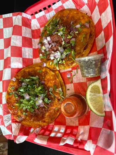 The image shows a serving of two tacos, likely birria tacos, presented on a red and white checkered paper liner within a red plastic basket. The tacos are topped with chopped red onions and cilantro. Accompaniments include a lime wedge, a small plastic container of green salsa, and another small plastic container of red salsa. The setting appears to be indoors, with a black surface visible in the periphery. The location is Columbus, United States of America.