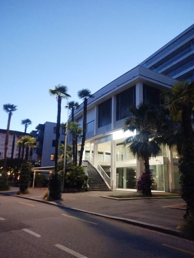 The image depicts a modern building with a glass facade and multiple floors, situated in the city of Muralto, Switzerland. The scene is set during twilight, indicated by the clear blue sky with subtle gradients and the artificial lighting emanating from the building. In front of the building, several palm trees stand tall, some with lush green fronds and others appearing more sparse. A paved road with distinct yellow and white lane markings runs in the foreground, suggesting it's a street or pathway. Adjacent to the road, a sidewalk or pedestrian area is visible, bordered by manicured greenery and more palm trees. A set of outdoor stairs leads up to the building's entrance, which is illuminated by a warm, white light. The overall impression is one of contemporary urban architecture integrated with a tropical or subtropical landscaping element. There are no people or vehicles visible in the frame.