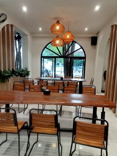 The image shows a cafe interior with a wooden table and chairs in the center of the room. The table is set with an empty plate and a napkin dispenser.  The table is made of dark wood and the chairs are black metal with wooden seats. There are three hanging lamps in the shape of round woven baskets.  The room is lit by overhead lighting. There is a large, arched window that looks out onto a street scene. The window is framed with black metal and has a decorative grille.  The room has white walls with wooden paneling on one side.  The location, based on the coordinates provided, is a cafe in Pati, Indonesia.