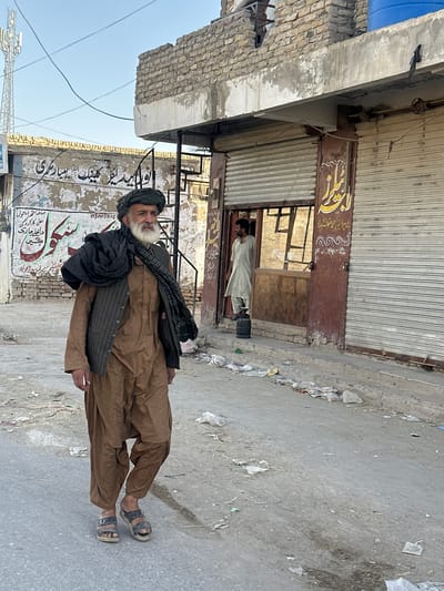 This image captures a street scene in Quetta City Tehsil, Pakistan. The primary subject is an older man standing in the foreground, wearing traditional Pakistani clothing: a brown shalwar kameez and a dark shawl draped over his shoulders. He has a white beard and is wearing a turban. He is looking towards the right side of the frame, with a neutral expression.

Behind him, there is a storefront with a metal roll-up shutter. A second man, wearing a light-colored shalwar kameez, is visible inside the shop, appearing to be engaged in some activity. The storefront has some signage with Arabic script, suggesting it might be a business or clinic. The street itself is paved and appears somewhat worn, with debris and litter scattered along the edges. The buildings in the background are made of brick and concrete, typical of urban Pakistani architecture. The lighting suggests it is daytime, likely late morning or early afternoon, given the clear sky. The overall impression is of a typical day in a busy commercial area.

Visible text in the image includes what appears to be business names or contact information in Arabic script. Some readable text in Urdu script can also be identified, including "پروپرایز محمد شرف شهداد" (Proprietor Muhammad Sharaf Shahdad), "میں ڈرایو کلینک بسیار سری" (in Drive Clinic Basiari Sari), and a phone number "03003892". Other legible phrases are "صاف ستھر اما حول" (Clean and tidy environment), "اعلی کارکردگی" (High performance), "داخلے جاری" (Admissions open), and "ال روڈ کوئٹہ" (Al Road Quetta).
