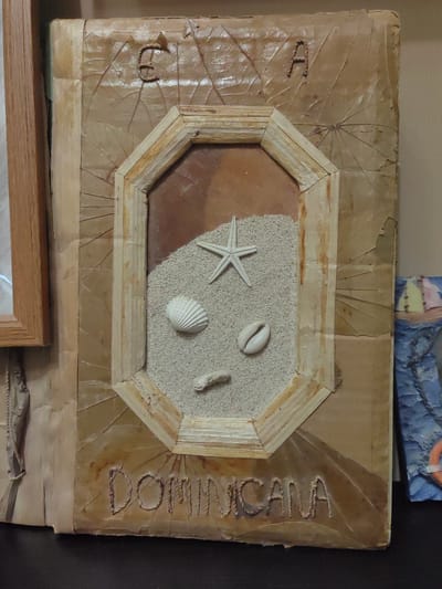 The image shows a framed piece of art with a beach theme. The frame is made of what appears to be dried leaves and straw. Inside the frame, there is sand, a starfish, and shells. The words "DOMINICANA" are carved into the bottom of the frame. The artwork is displayed on a dark surface. There is another framed object on the left and a small boat-themed decoration on the right. The image does not provide any information about the location in the city of Ostafievo, Russia.