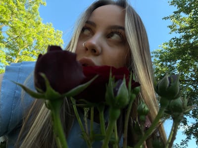 An upward-facing, low-angle shot captures a young person holding dark red roses against a clear blue sky, likely in Novopokrovskaya, Russia. The individual's face is seen from below, with their eyes looking directly down towards the camera. Long, light brown hair frames the face, which features a nose ring. The roses, some still in bud form and others partially open, are held above the face, with their green stems and leaves prominent in the foreground. A glimpse of a light blue garment, possibly a denim shirt or jacket, is visible near the top right. In the background, lush green tree foliage contrasts with the vibrant blue sky, suggesting an outdoor setting during daytime with clear weather. No visible text is present.