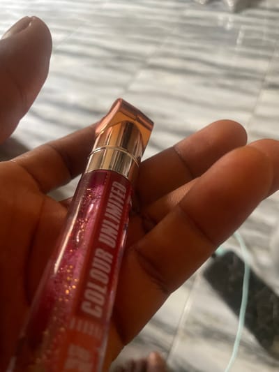 A hand is holding a tube of lip gloss. The lip gloss is red with gold glitter. The tube is clear with a gold cap and a gold band around the top. The word "COLOUR UNLIMITED" is written on the side of the tube in white letters. The background is a marble surface. The image is cropped to show the hand and the lip gloss.