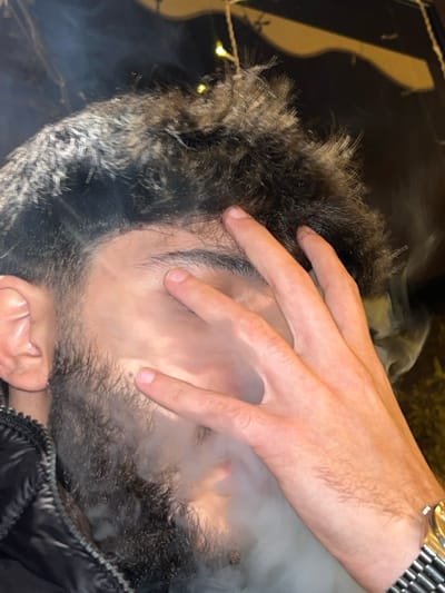 A close-up, angled shot shows a person with dark hair and a beard, their face obscured by a hand and a cloud of smoke. The hand, adorned with a gold wristwatch, rests over the person's eyes, partially concealing their face. The smoke swirls around their face and the hand. A dark jacket is visible along the bottom right edge. A blurred background hints at some lights.